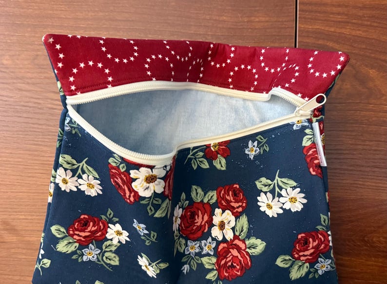 K&ouml;nnte beinhalten: Eine Rei&szlig;verschlusstasche mit floralem Muster. Der marineblaue Stoff ist mit roten Rosen und wei&szlig;en G&auml;nsebl&uuml;mchen verziert. Der obere Rand ist tiefrot mit wei&szlig;en Sternen. Der Rei&szlig;verschluss ist beige.