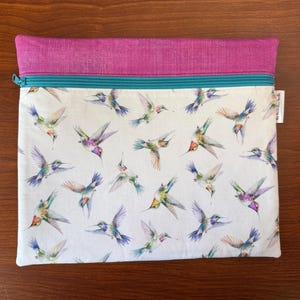 May include: A rectangular zippered pouch with a white background and a pattern of colorful hummingbirds in flight. The top section is a solid magenta color, and the zipper is teal. A small white tag is attached to the side.