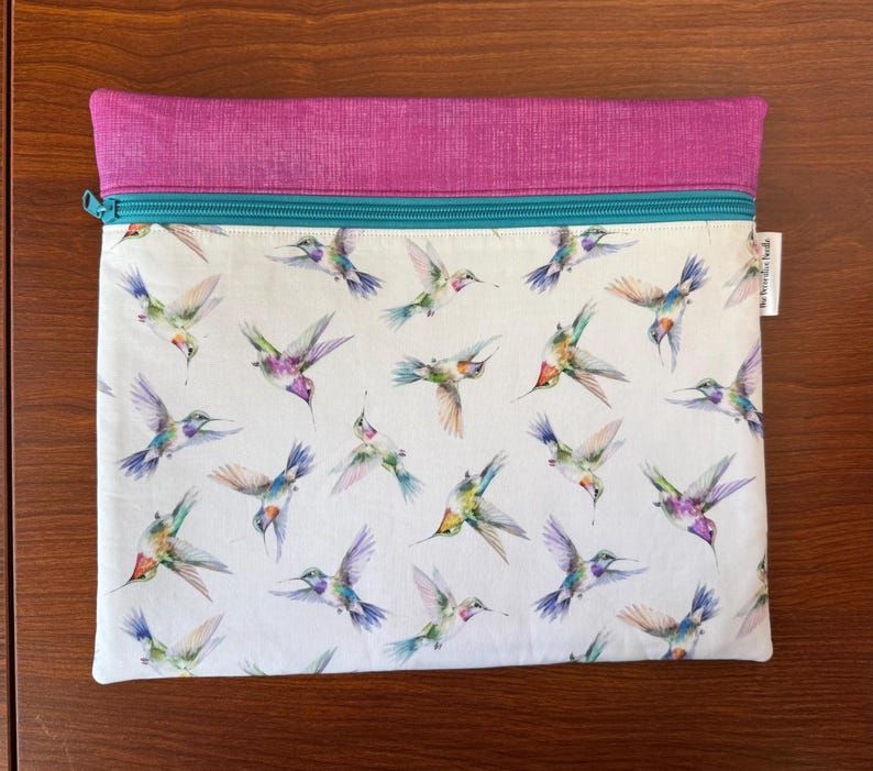 May include: A rectangular zippered pouch with a white background and a pattern of colorful hummingbirds in flight. The top section is a solid magenta color, and the zipper is teal. A small white tag is attached to the side.