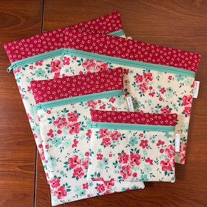 Puede incluir: Un conjunto de cinco bolsas de tela con cremallera de diferentes tama&ntilde;os. Las bolsas presentan un estampado floral crema con flores rosas y azules. Cada bolsa tiene un borde superior rojo y una cremallera turquesa.