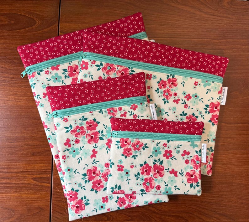 Puede incluir: Un conjunto de cinco bolsas de tela con cremallera de diferentes tama&ntilde;os. Las bolsas presentan un estampado floral crema con flores rosas y azules. Cada bolsa tiene un borde superior rojo y una cremallera turquesa.