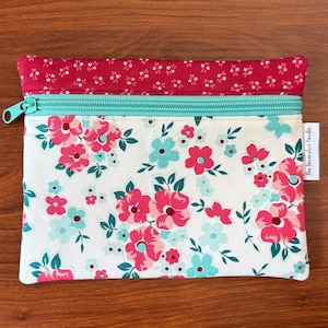 Puede incluir: Una bolsa rectangular con estampado floral con cremallera turquesa y parte superior floral roja. La bolsa presenta un fondo blanco con flores rosas y turquesas y hojas verdes. El texto "the decorative needle" est&aacute; impreso en una peque&ntilde;a etiqueta blanca.