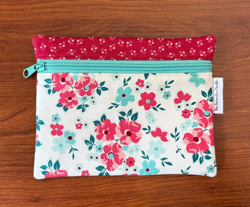 Puede incluir: Una bolsa rectangular con estampado floral con cremallera turquesa y parte superior floral roja. La bolsa presenta un fondo blanco con flores rosas y turquesas y hojas verdes. El texto "the decorative needle" est&aacute; impreso en una peque&ntilde;a etiqueta blanca.