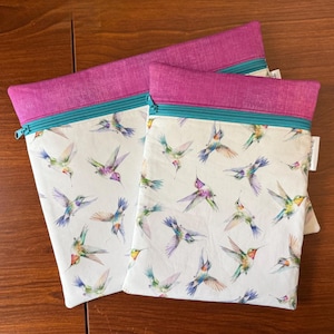 May include: Two zippered pouches with a white background and a colorful hummingbird pattern. The top of each pouch is a vibrant magenta, with a teal zipper. The pouches are made of fabric.