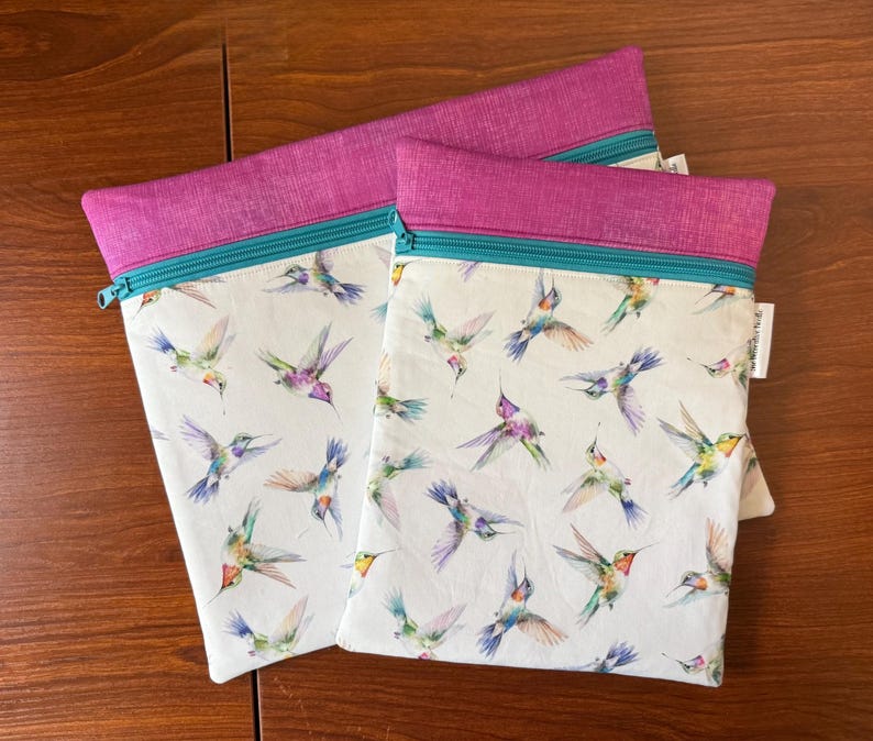 May include: Two zippered pouches with a white background and a colorful hummingbird pattern. The top of each pouch is a vibrant magenta, with a teal zipper. The pouches are made of fabric.