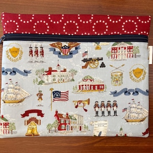 Op de afbeelding: Een rechthoekig stoffen etui met een rood en wit sterrenpatroon aan de bovenkant en een lichtblauwe achtergrond. De etui heeft een ritssluiting en is versierd met patriottische illustraties, waaronder gebouwen, vlaggen en soldaten.