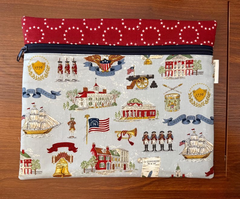 Op de afbeelding: Een rechthoekig stoffen etui met een rood en wit sterrenpatroon aan de bovenkant en een lichtblauwe achtergrond. De etui heeft een ritssluiting en is versierd met patriottische illustraties, waaronder gebouwen, vlaggen en soldaten.