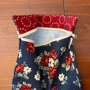 K&ouml;nnte beinhalten: Eine marineblaue Rei&szlig;verschlusstasche mit einem Blumenmuster aus roten Rosen und wei&szlig;en G&auml;nsebl&uuml;mchen. Der obere Rand ist mit einem roten Stoff mit einem wei&szlig;en Sternmuster gef&uuml;ttert. Der Rei&szlig;verschluss ist beige.