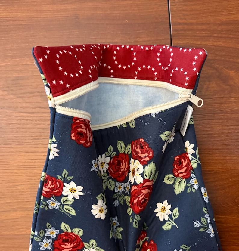 K&ouml;nnte beinhalten: Eine marineblaue Rei&szlig;verschlusstasche mit einem Blumenmuster aus roten Rosen und wei&szlig;en G&auml;nsebl&uuml;mchen. Der obere Rand ist mit einem roten Stoff mit einem wei&szlig;en Sternmuster gef&uuml;ttert. Der Rei&szlig;verschluss ist beige.