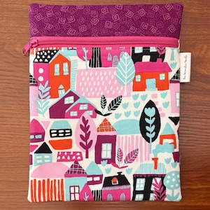 Puede incluir: Un estuche rectangular de tela con cremallera que presenta un patr&oacute;n colorido de casas y &aacute;rboles en tonos naranja, rosa, morado, negro y azul claro. La secci&oacute;n superior es de tela morada s&oacute;lida con un patr&oacute;n geom&eacute;trico y una cremallera rosa.