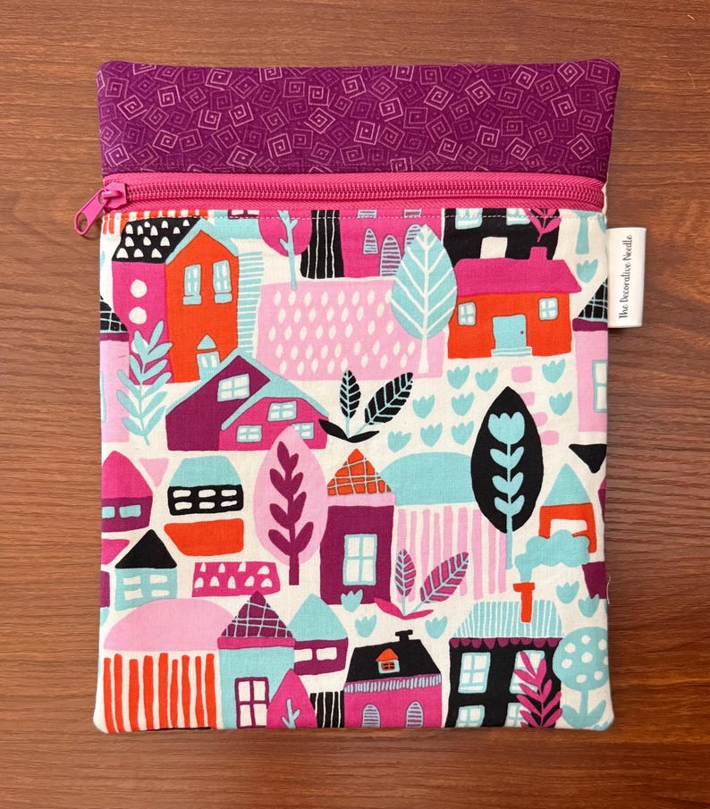 Puede incluir: Un estuche rectangular de tela con cremallera que presenta un patr&oacute;n colorido de casas y &aacute;rboles en tonos naranja, rosa, morado, negro y azul claro. La secci&oacute;n superior es de tela morada s&oacute;lida con un patr&oacute;n geom&eacute;trico y una cremallera rosa.