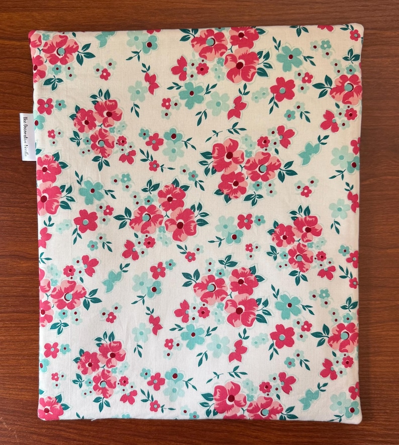 Puede incluir: Un art&iacute;culo de tela rectangular con estampado floral. La tela presenta un fondo blanco con un patr&oacute;n repetido de flores rosas y turquesas y hojas verdes. El art&iacute;culo tiene una etiqueta que dice "The Decorative Handi".