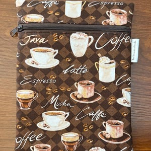 May include: A rectangular, zippered pouch with a brown coffee-themed pattern. The design features coffee cups, coffee beans, and coffee-related words like "espresso" and "latte" on a brown and tan checkered background. The zipper is dark brown.
