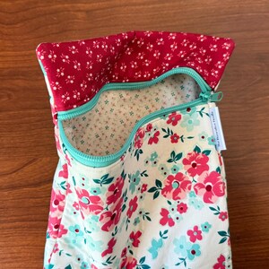 Puede incluir: Una bolsa de tela floral con cremallera turquesa. La bolsa presenta un fondo blanco con flores rosas y turquesas. El forro interior es un peque&ntilde;o estampado floral sobre un fondo crema. El borde superior es de tela roja con detalles florales blancos.