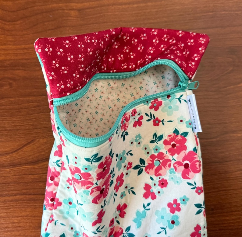 Puede incluir: Una bolsa de tela floral con cremallera turquesa. La bolsa presenta un fondo blanco con flores rosas y turquesas. El forro interior es un peque&ntilde;o estampado floral sobre un fondo crema. El borde superior es de tela roja con detalles florales blancos.