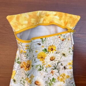 K&ouml;nnte beinhalten: Ein Stoffbeutel mit Rei&szlig;verschluss und einem Blumenmuster aus gelben und wei&szlig;en G&auml;nsebl&uuml;mchen, gr&uuml;nen Bl&auml;ttern und kleinen Bienen auf einem Hintergrund mit kursivem Text. Der Beutel hat einen gelben Rei&szlig;verschluss und ein gelbes Innenfutter.