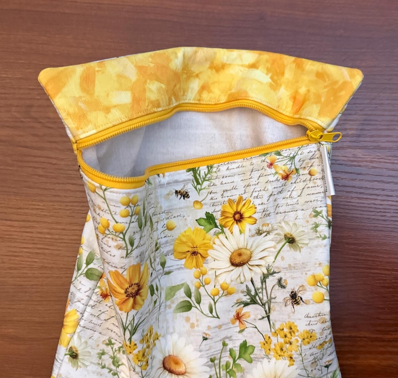 K&ouml;nnte beinhalten: Ein Stoffbeutel mit Rei&szlig;verschluss und einem Blumenmuster aus gelben und wei&szlig;en G&auml;nsebl&uuml;mchen, gr&uuml;nen Bl&auml;ttern und kleinen Bienen auf einem Hintergrund mit kursivem Text. Der Beutel hat einen gelben Rei&szlig;verschluss und ein gelbes Innenfutter.