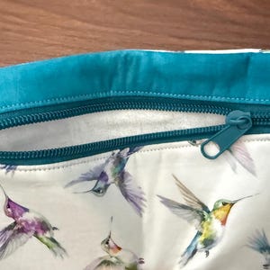 May include: A white zippered pouch featuring a hummingbird print in shades of purple, green, and yellow. The top edge and zipper are teal. The pouch has a small white tag that reads "The Decorative Needle".