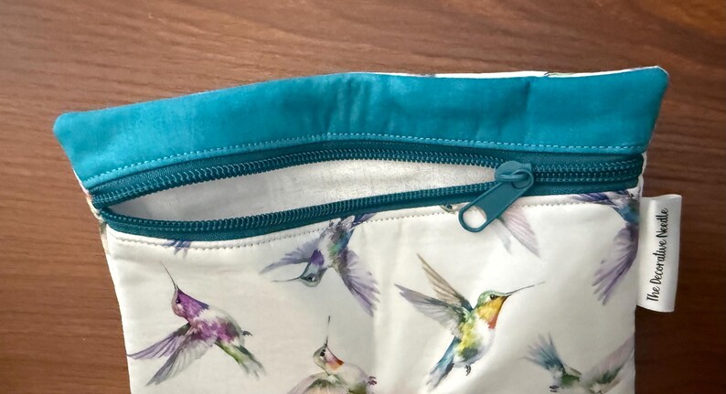 May include: A white zippered pouch featuring a hummingbird print in shades of purple, green, and yellow. The top edge and zipper are teal. The pouch has a small white tag that reads "The Decorative Needle".