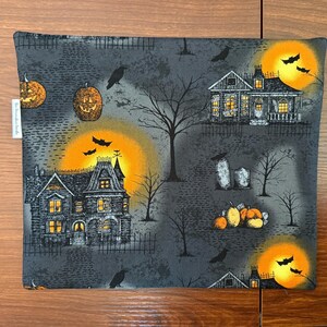 May include: A square fabric pouch featuring a dark gray Halloween print with haunted houses, glowing orange full moons, black bats, bare trees, jack-o'-lanterns, and tombstones. A small white fabric tag is attached to the left side edge.