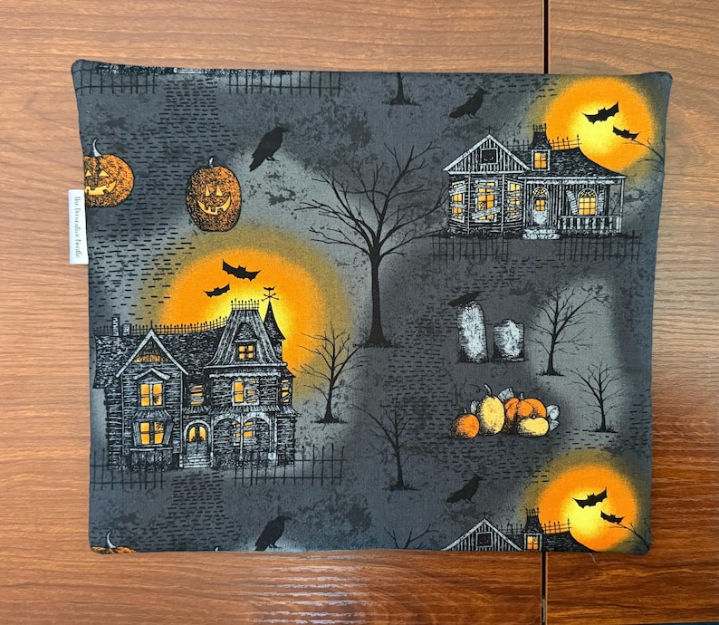 May include: A square fabric pouch featuring a dark gray Halloween print with haunted houses, glowing orange full moons, black bats, bare trees, jack-o'-lanterns, and tombstones. A small white fabric tag is attached to the left side edge.