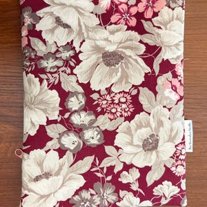 May include: A rectangular fabric pouch featuring a deep burgundy background with a large-scale floral pattern in shades of cream, light pink, and muted brown. A small white fabric tag on the side displays the text The Decorative Needle.