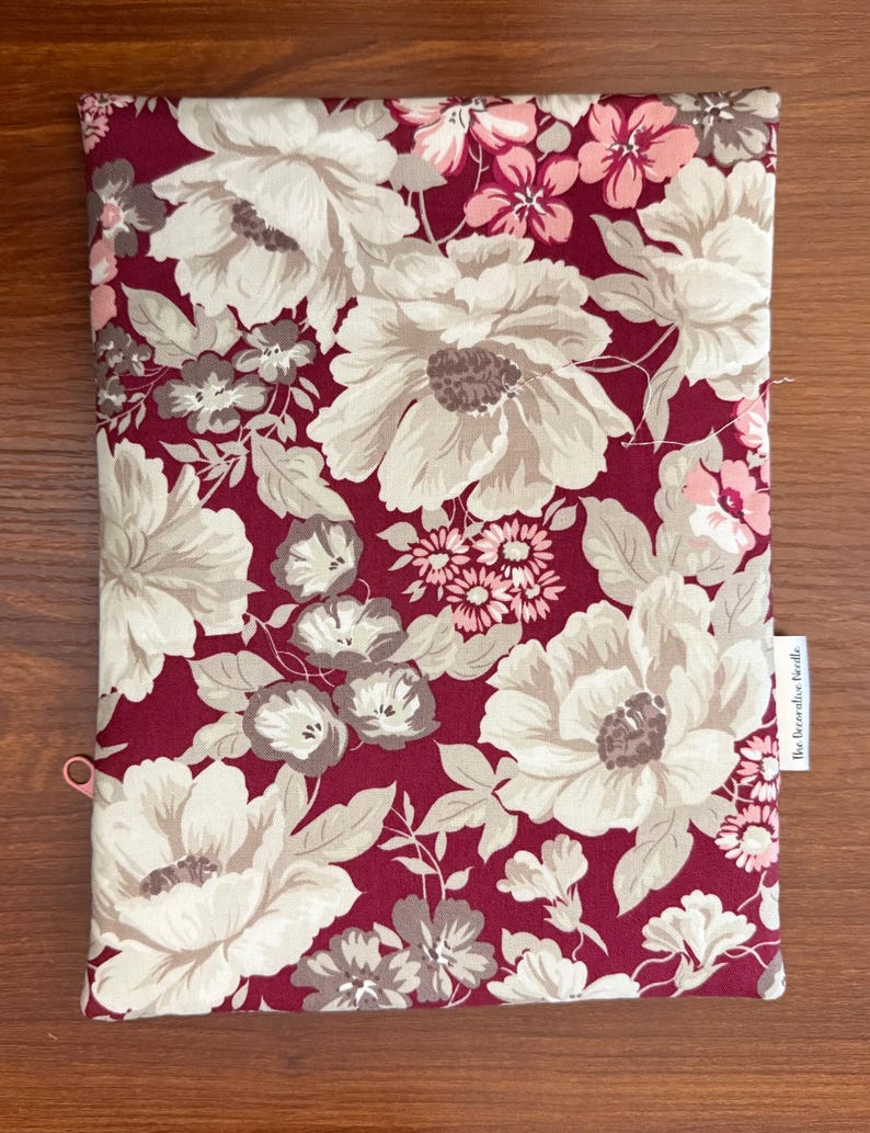 May include: A rectangular fabric pouch featuring a deep burgundy background with a large-scale floral pattern in shades of cream, light pink, and muted brown. A small white fabric tag on the side displays the text The Decorative Needle.