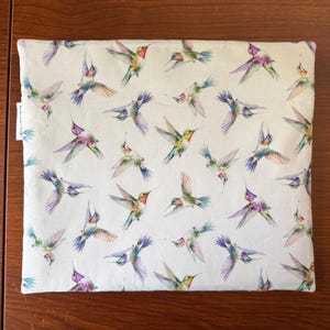 May include: A white fabric item with a repeating pattern of colorful hummingbirds in flight. The birds feature shades of green, purple, blue, and red. The item is rectangular and appears to be a pillowcase or similar textile.