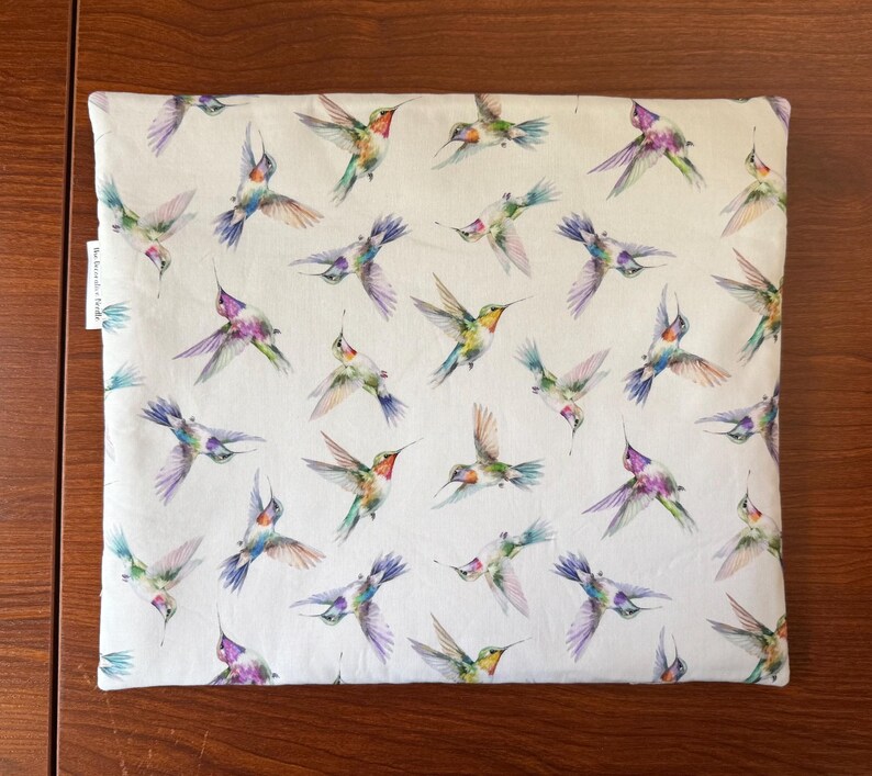 May include: A white fabric item with a repeating pattern of colorful hummingbirds in flight. The birds feature shades of green, purple, blue, and red. The item is rectangular and appears to be a pillowcase or similar textile.