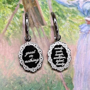 May include: A pair of silver hoop earrings with a charm attached to each. The charm on the left earring has the words "great or nothing" inscribed on it. The charm on the right earring has the words "minds, souls, hearts, ambition, talent, beauty" inscribed on it.