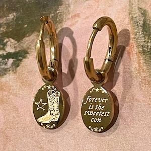 Puede incluir: Un par de pendientes de aro de oro con un dije colgando de cada uno. Un dije presenta una bota de vaquero con una estrella, el otro dije dice "forever is the sweetest con".