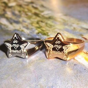 Puede incluir: Dos anillos de oro y plata con un diseño en forma de estrella. La estrella tiene las palabras "A in my mind" grabadas en ella. Los anillos están sobre una superficie con un patrón azul y blanco.