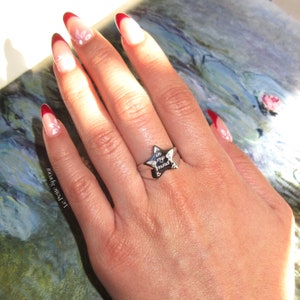 Pu&ograve; includere: Un anello d'argento con un design a forma di stella. La stella ha le parole "in my mind" incise. L'anello &egrave; indossato su una mano con smalto rosso e bianco.