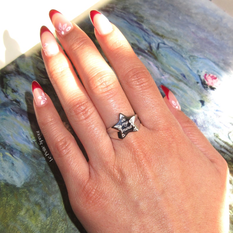 Pu&ograve; includere: Un anello d'argento con un design a forma di stella. La stella ha le parole "in my mind" incise. L'anello &egrave; indossato su una mano con smalto rosso e bianco.