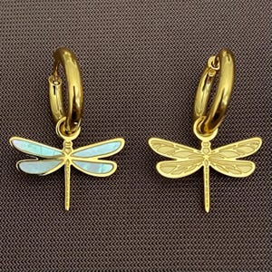 May include: A pair of gold-coloured hoop earrings featuring dragonfly charms. One dragonfly charm has iridescent blue-green wings, the other has gold-coloured wings. The earrings are displayed on a brown surface.