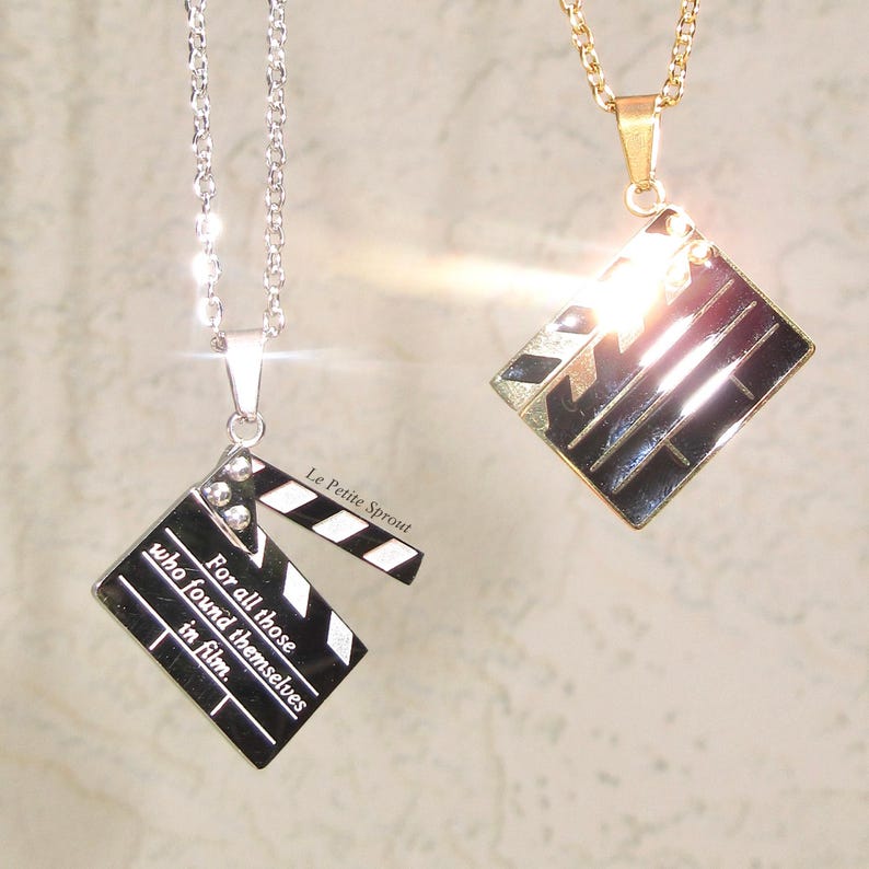 May include: Two silver and gold necklaces with a clapperboard pendant. The silver necklace has the text "For all those who found themselves in film." The gold necklace has a shiny, reflective surface.