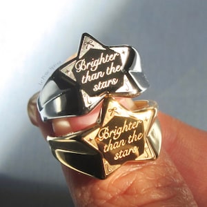 Könnte beinhalten: Zwei goldene sternförmige Ringe mit der Inschrift "Brighter than the stars" auf jedem. Die Ringe sind übereinander gestapelt.