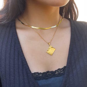 May include: A gold chain necklace with a square pendant. The pendant has a textured surface and is engraved with the words "Less is More".