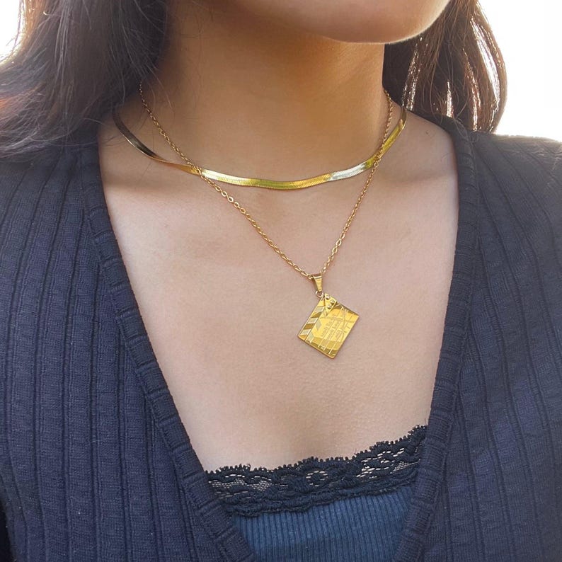 May include: A gold chain necklace with a square pendant. The pendant has a textured surface and is engraved with the words "Less is More".