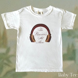 May include: White baby tee with a brown and tan graphic of headphones and the text "The Pleasure's All Mine". The shirt has short sleeves and a crew neck. The words "Baby Tee" are at the bottom.