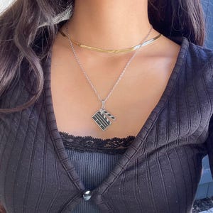 May include: A silver chain necklace with a silver pendant shaped like a clapperboard. The pendant is black and white and has a silver chain attached to it.