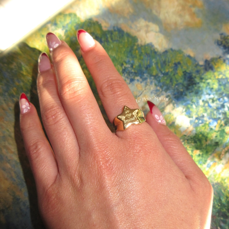 Pu&ograve; includere: Anello dorato a forma di stella con le parole "in my mind" incise sulla superficie. L'anello &egrave; indossato su un dito.
