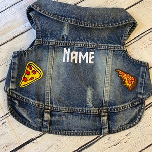 May include: A blue denim vest with a white embroidered name and two pizza slice patches. The patches are yellow, red, and orange.