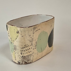 May include: A white ceramic planter with a rectangular shape and a distressed finish. The planter has a light green and black abstract design.