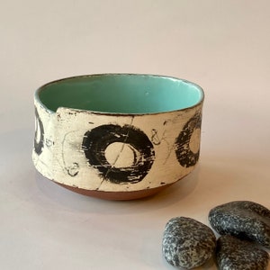 May include: A small, white ceramic bowl with a light blue interior. The bowl has a unique design of black circles with white centers. The bowl is sitting on a white surface.