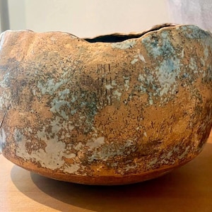 May include: A round ceramic bowl with a textured, uneven rim. The exterior features a mottled finish with earthy brown, copper, and oxidized teal green tones. A small, vertical stamped mark is visible on the side of the bowl's curved surface.