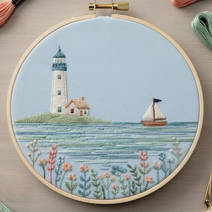 Può includere: Opera d'arte ricamata in un cerchio di legno raffigurante una scena costiera. Un faro bianco e una casetta si trovano su un'isola verde. Una barca a vela con una bandiera blu naviga sull'acqua blu. Fiori e vegetazione colorati in primo piano.