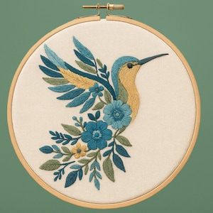 May include: Embroidered hummingbird design in a wooden hoop. The bird features blue, yellow, and green thread, surrounded by floral and leaf details. The background is a cream-colored fabric.