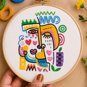 May include: Hand-embroidered abstract face design on white fabric stretched in a wooden hoop. The artwork features bold lines and shapes in yellow, blue, pink, and green. The text "Hand Embroidery PDF Patterns by Zazarita" is at the bottom.