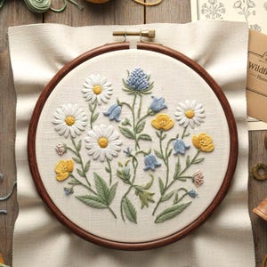 Pode incluir: Um bastidor de bordado detalhado com um design floral com margaridas brancas, botões-de-ouro amarelos e flores de campainha azuis. O bordado é feito sobre um tecido creme, emoldurado por um aro de madeira escura.
